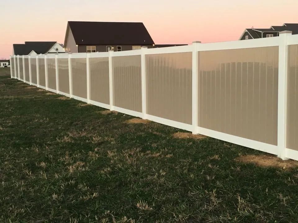 White vinyl privacy fence in a grassy yard, houses in background, dusk sky.
