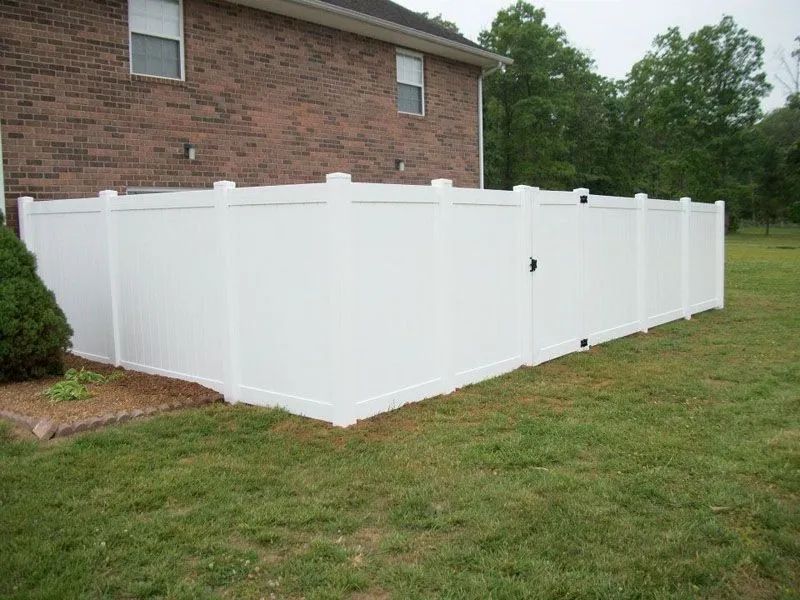 White vinyl fence encloses a backyard with a gate next to a brick building on a grassy lawn.