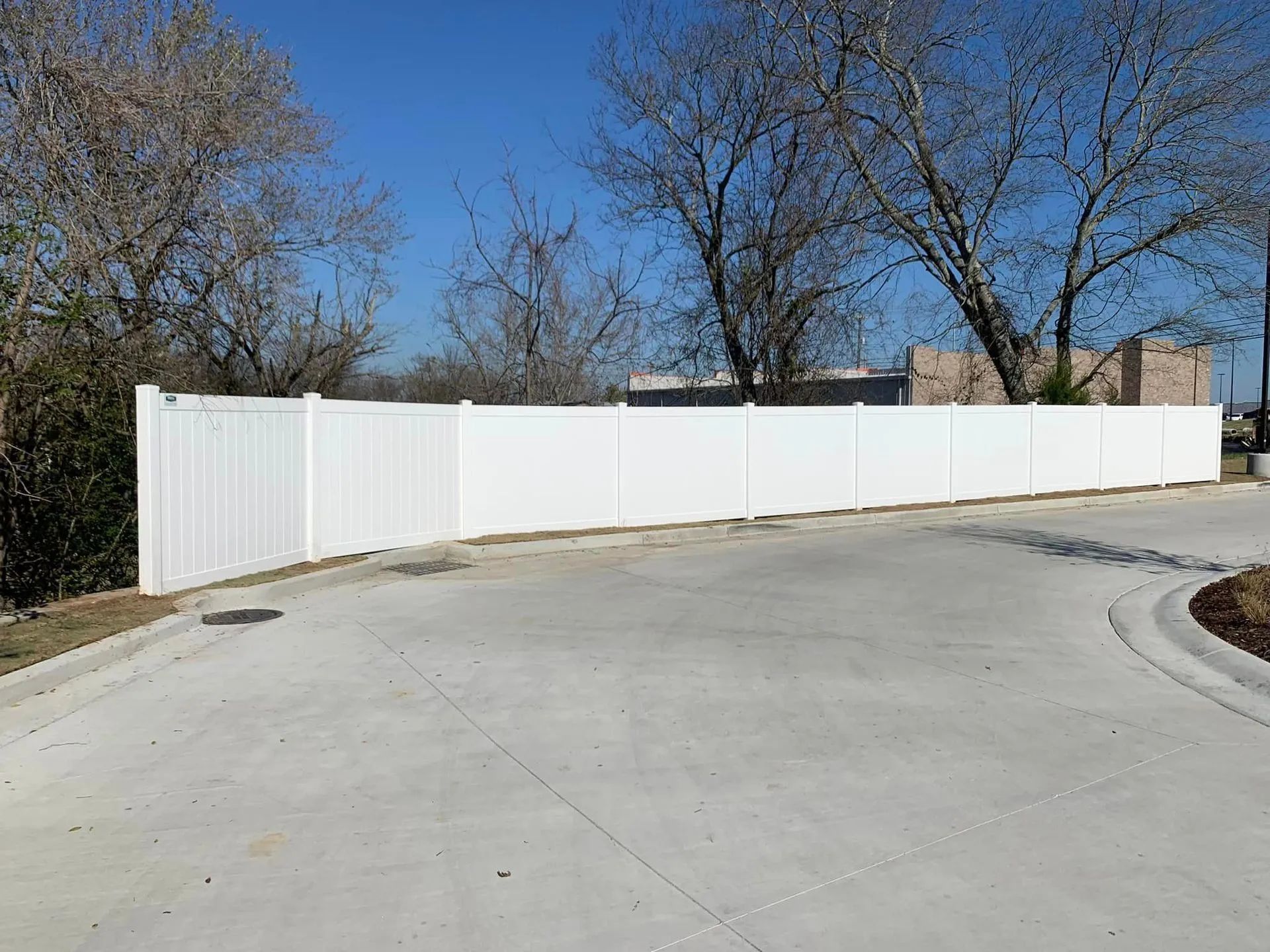 White vinyl fence bordering a concrete driveway and sidewalk, under a blue sky, with bare trees in the background.