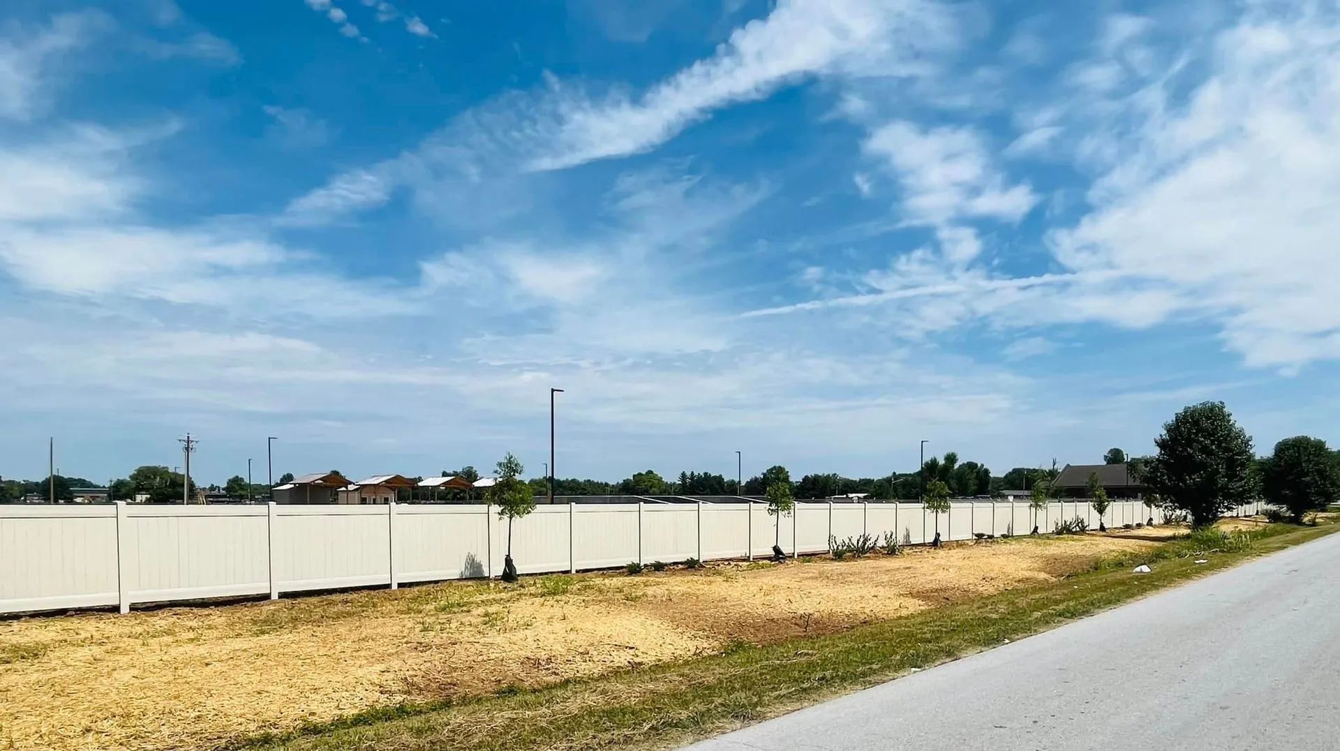 White fence borders a field under a blue sky with streaky clouds; a road is in the foreground.
