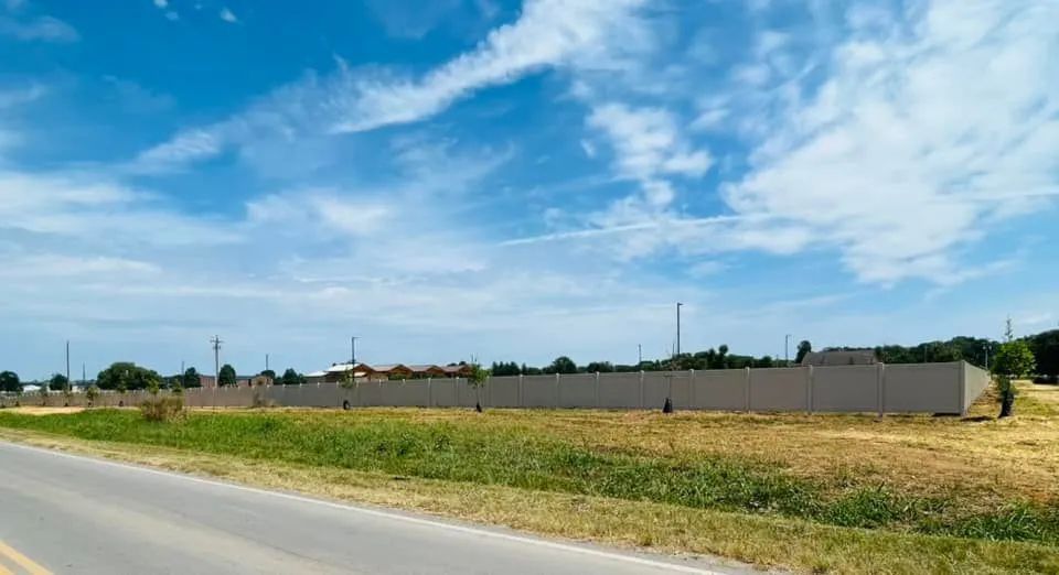 A long concrete wall along a field with a road and a blue sky with clouds.