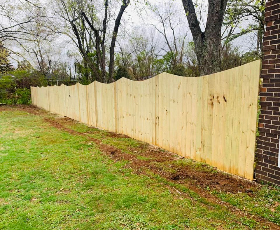 New, unfinished wooden fence along a grassy yard, with a wavy top. Brown dirt path adjacent.