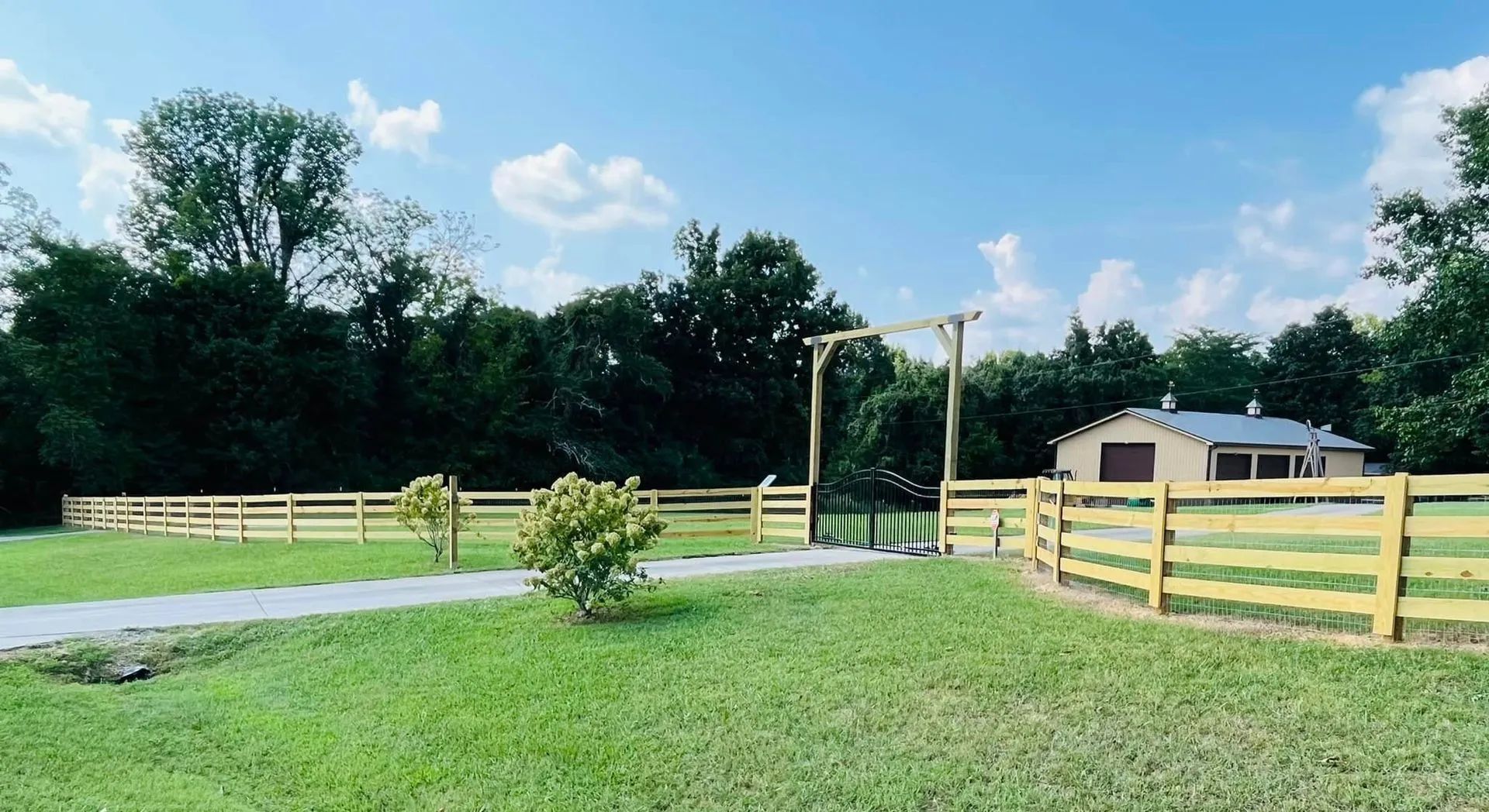 Wooden fence surrounds a grassy field with an entrance archway; a small building is visible in the background under a blue sky.