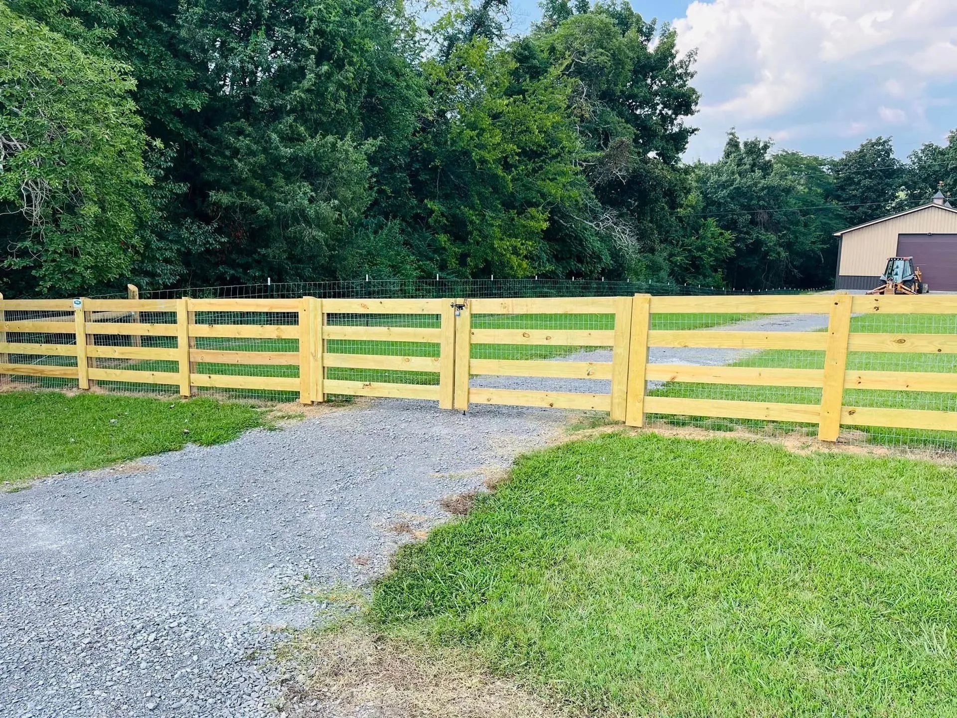Wooden fence with gate in front of a gravel driveway and grassy area, trees in the background.