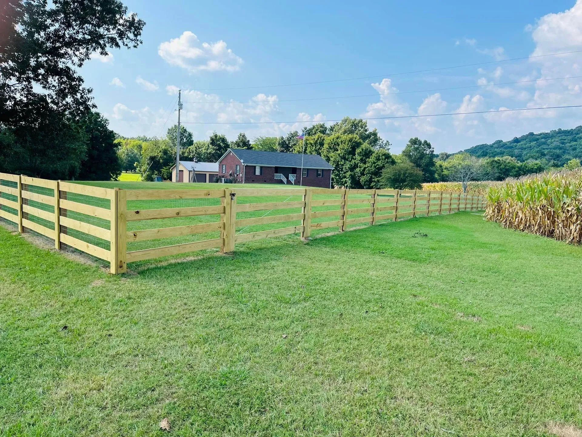 Wooden fence bordering a grassy field, house in the background, blue sky.
