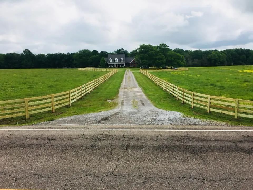 Long gravel driveway leading to a house, flanked by wooden fences, green field, and trees. Overcast sky.