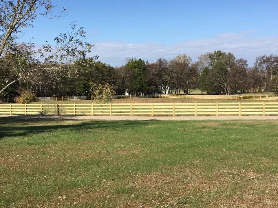 A long wooden fence in a grassy field, trees in the background, blue sky.