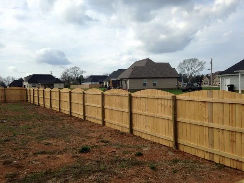 Wooden fence in a residential area, with arched tops, separating yards under a cloudy sky.