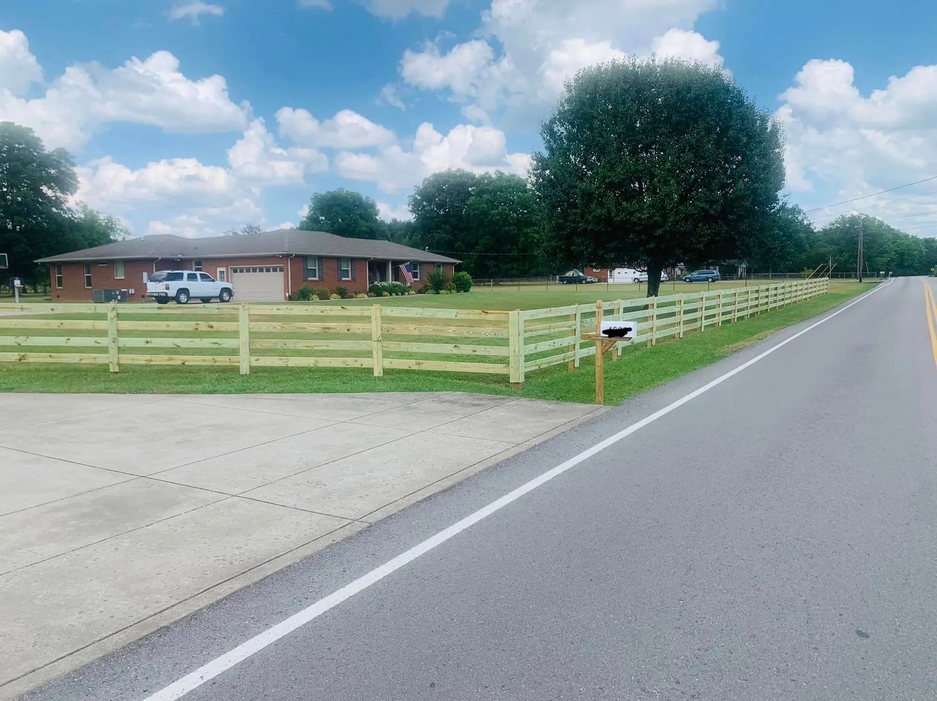 A two-story brick house with a white truck, a wooden fence, and a mailbox next to a road under a blue sky.