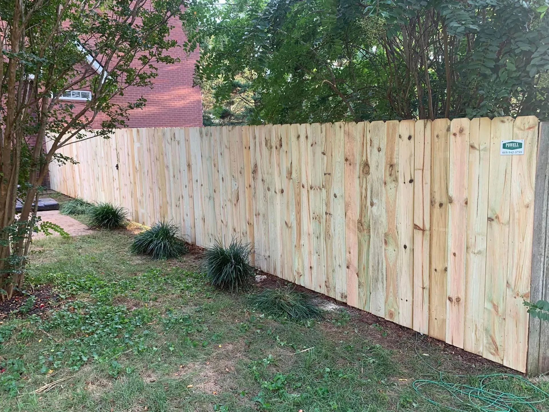 Wooden fence in a yard with grass and small bushes. Trees and a brick building are in the background.
