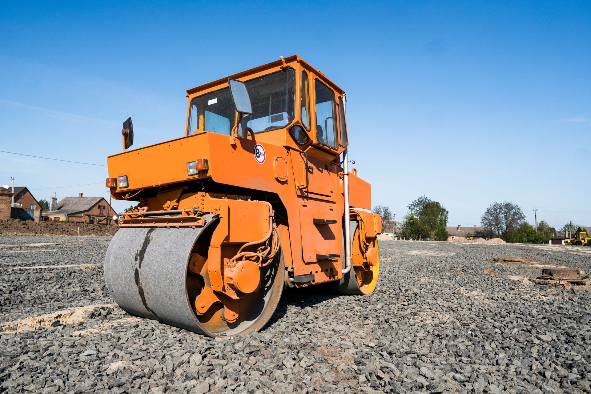 A bright orange industrial road roller parked on a gravel construction site under a clear blue sky.