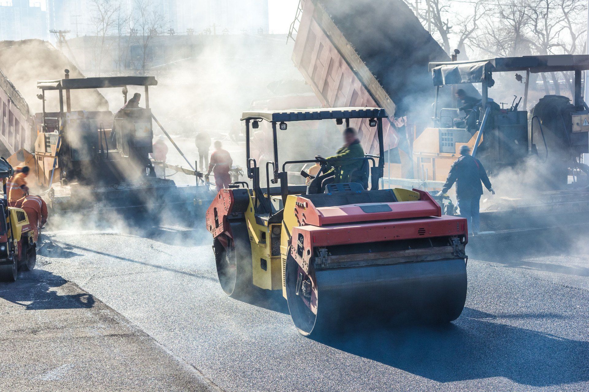 A road roller compacts freshly laid asphalt on a construction site, surrounded by steam and heavy equipment.