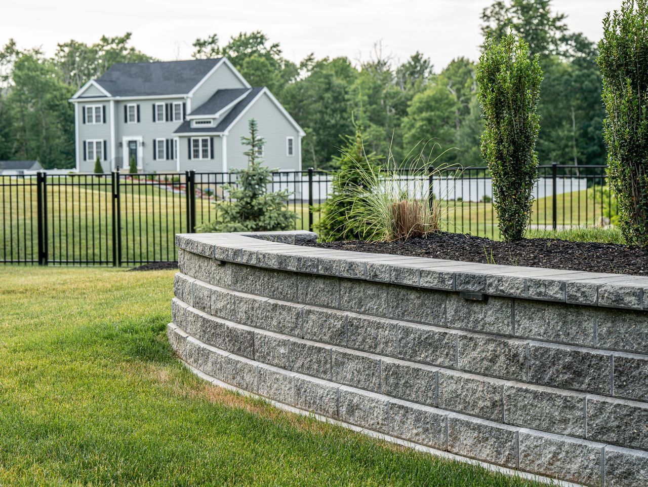 Curved stone retaining wall in a grassy yard with a black fence and a gray house in the background