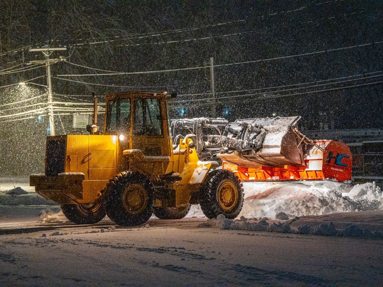Yellow snowplow clearing snow at night in a snowy street, with a red vehicle behind it.