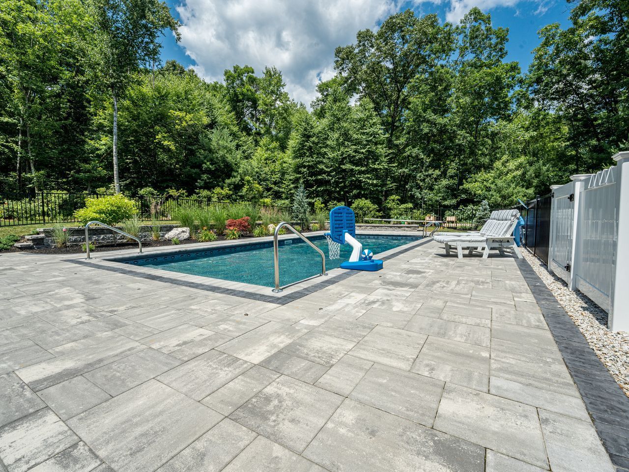 Backyard pool with turquoise water, blue slide, and patio chairs beside a white fence and trees