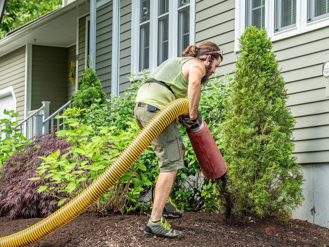 Person using a leaf blower to clear shrubs beside a house with green siding.