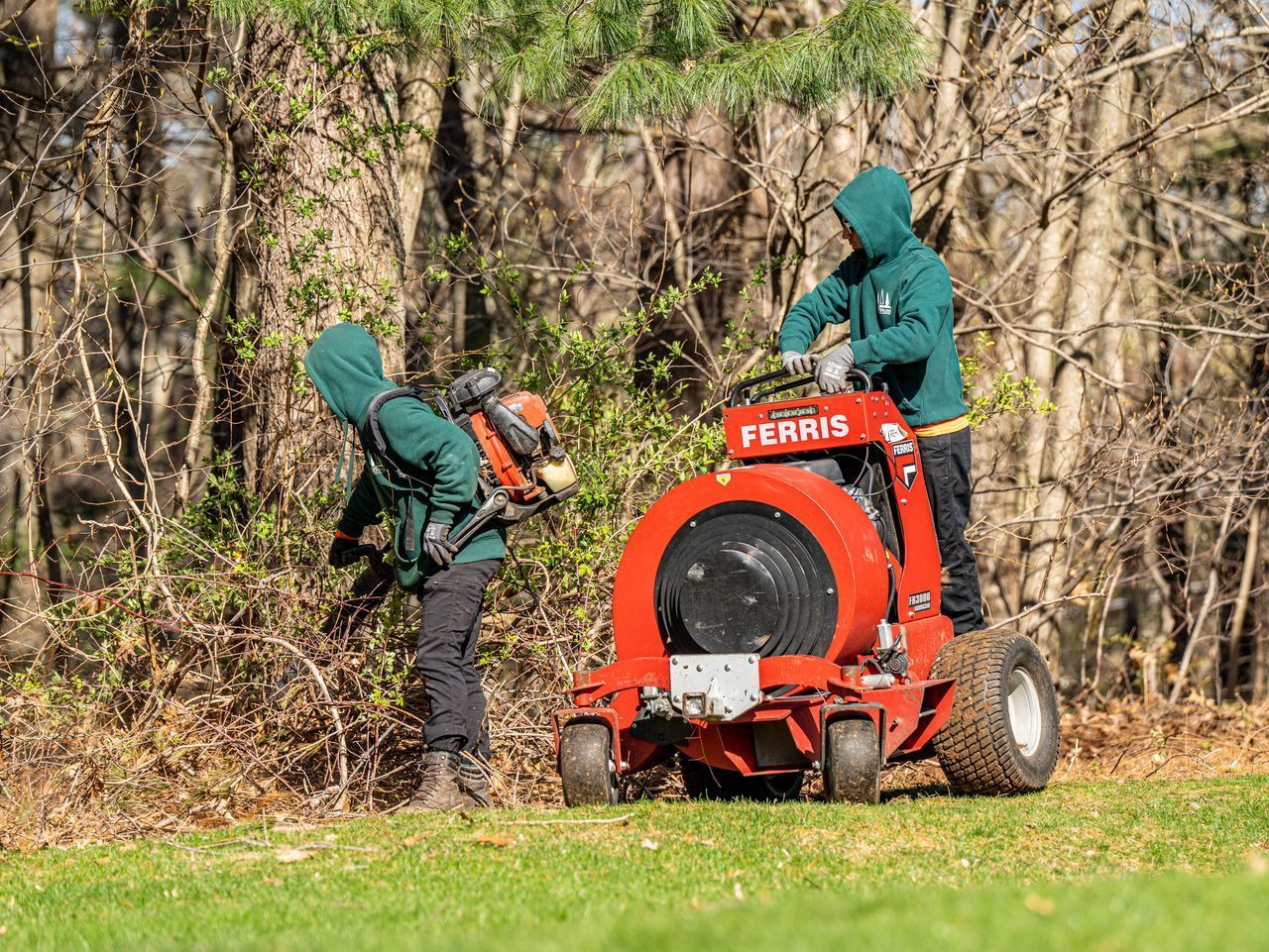 Two workers in green jackets operating a red Ferris machine on a grassy edge by trees.