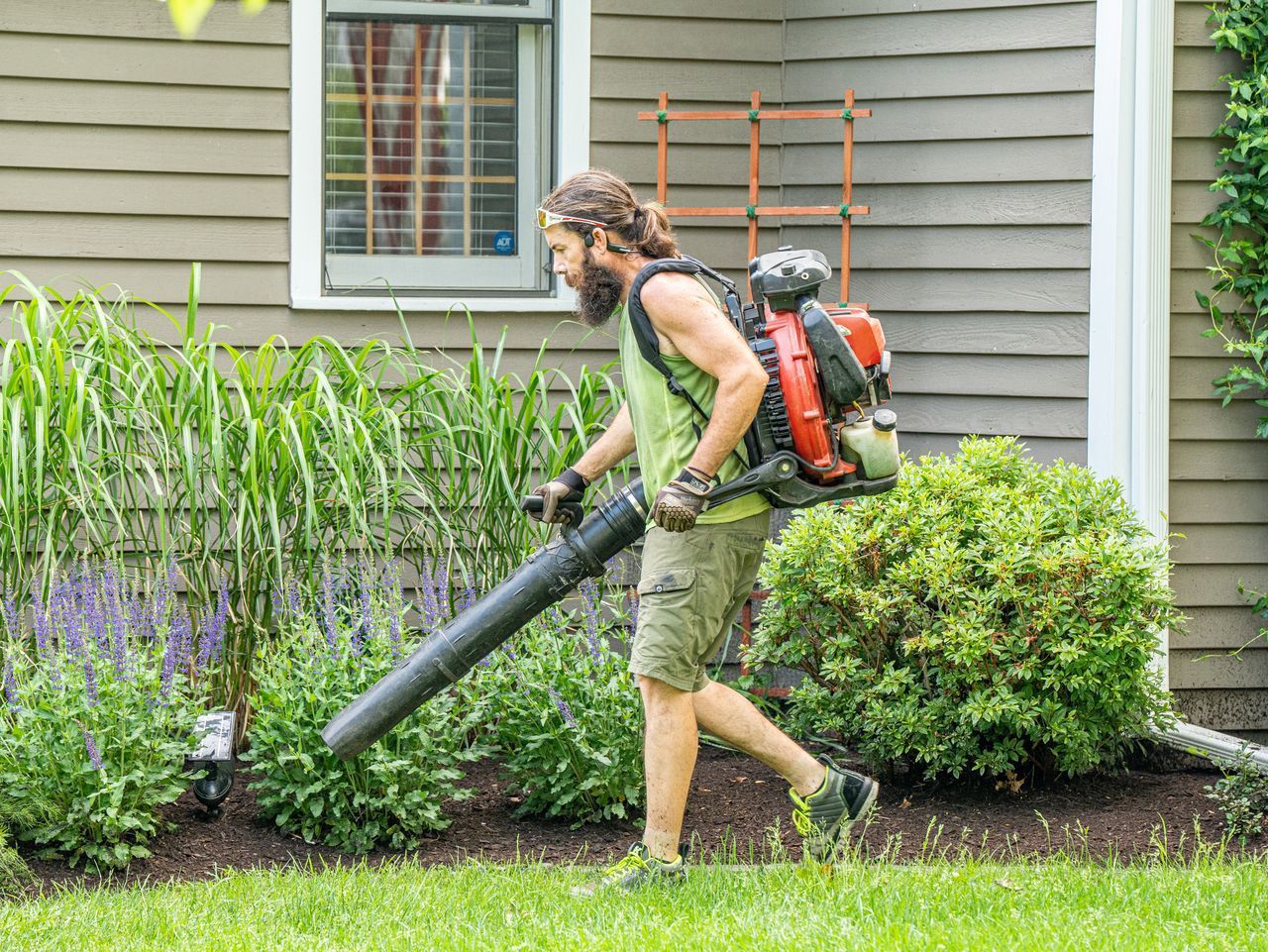 Person using a leaf blower beside a house, clearing grass and plants in a garden.