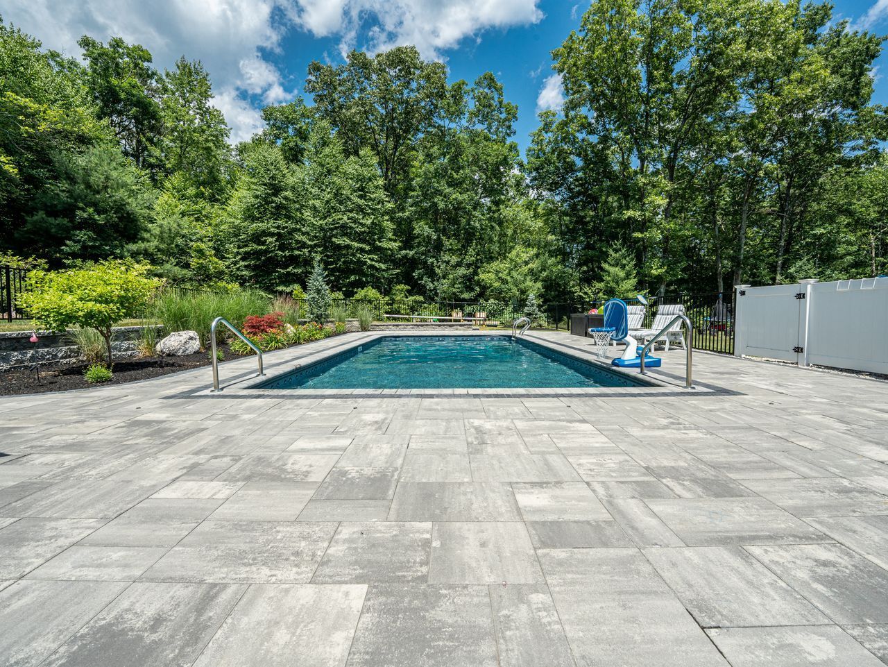 Backyard patio with a rectangular swimming pool, lounge chairs, and trees under a sunny sky