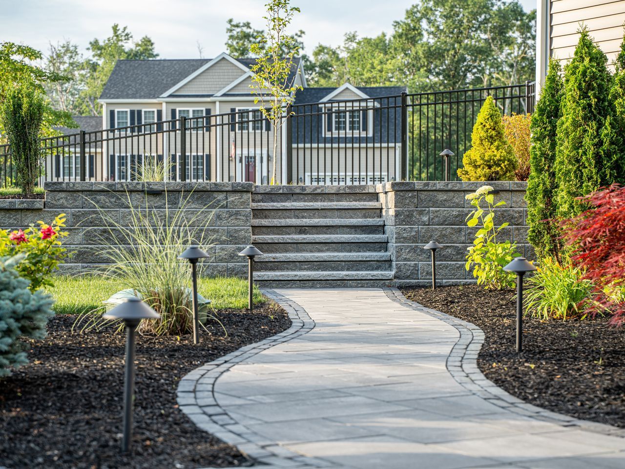 Curved garden path leading to stone steps, black fence, and a small house with landscaped shrubs and trees.