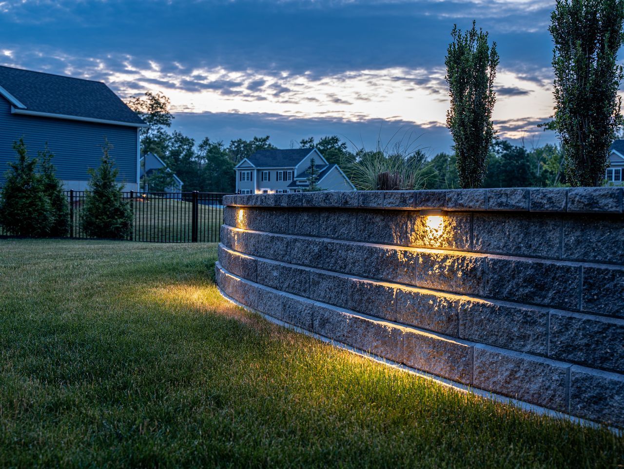Backyard retaining wall with warm lights at dusk, beside a grassy lawn and suburban houses.