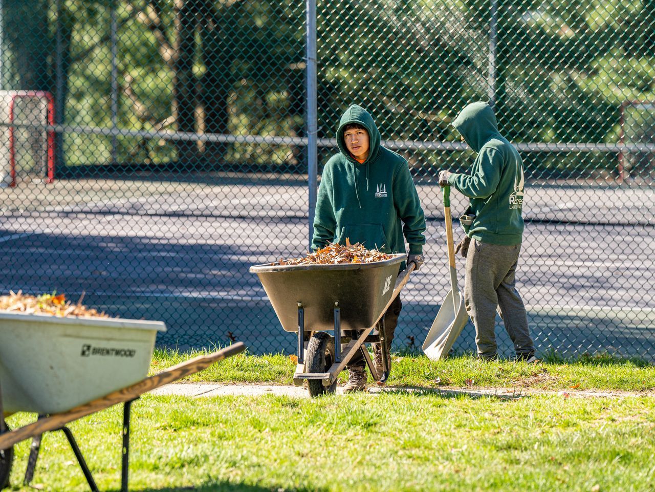 Two people in green hoodies rake leaves into a wheelbarrow by a chain-link fence in a park.