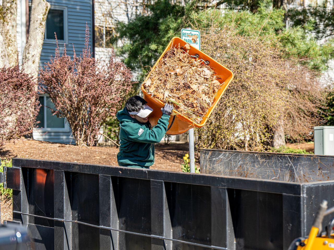 Person tipping a leaf-filled orange bin into a large black dumpster outdoors