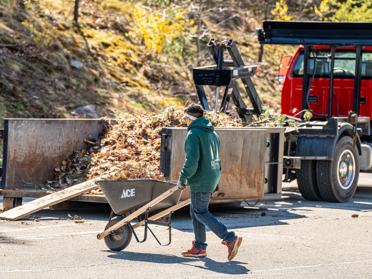Person in green jacket pushing a wheelbarrow of leaves beside a red truck unloading debris