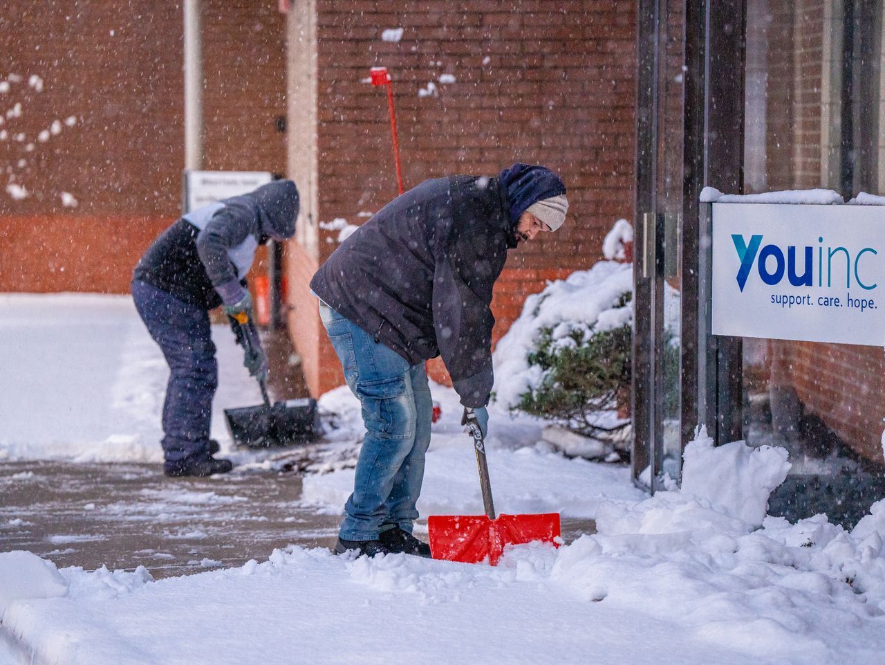 Two people shoveling snow outside a brick building, with a red shovel and falling snow.