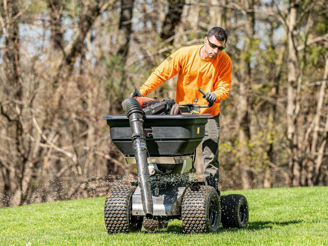 Person in an orange shirt spreading fertilizer on a lawn with a push spreader