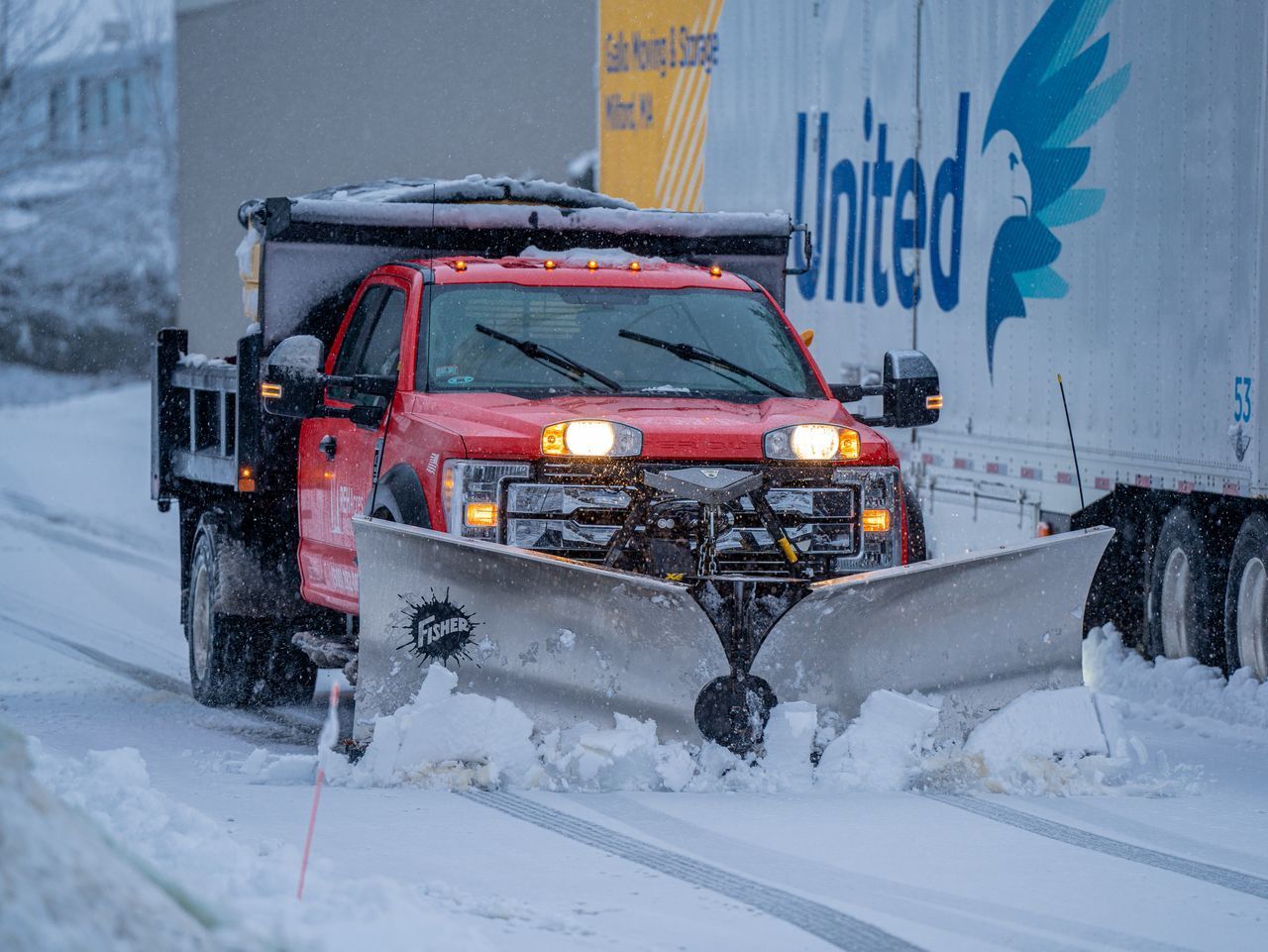 Red pickup truck plowing a snow-covered road in heavy snowfall, with a large white trailer beside it.