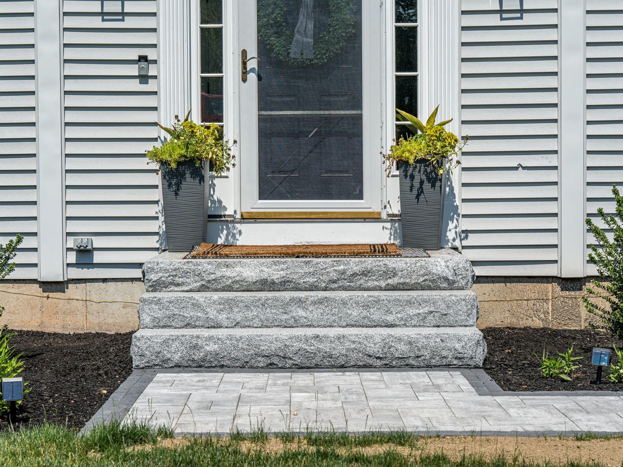 Front entry steps to a house with gray stone, white siding, and yellow flowers in planters