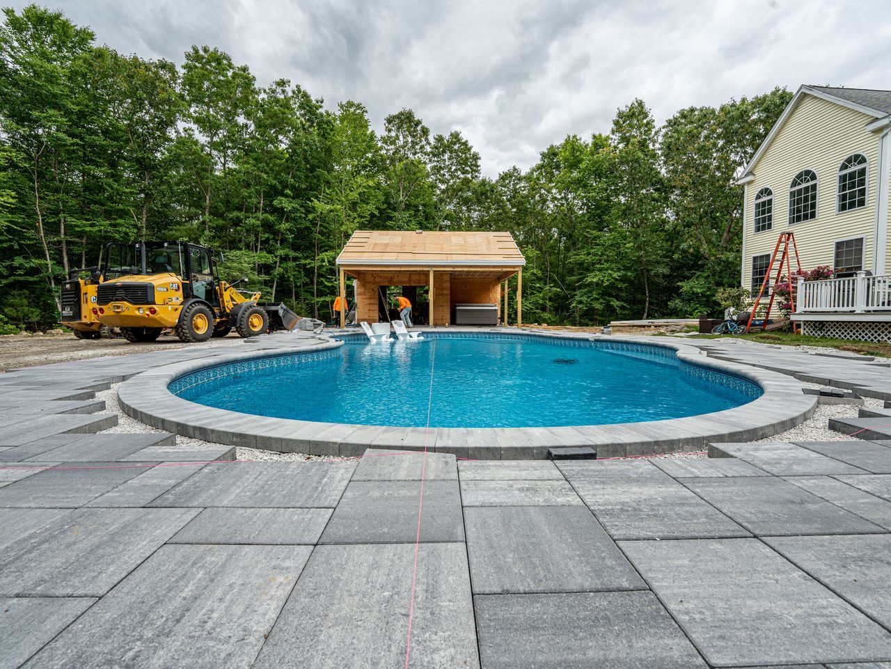 Backyard with round blue pool, stone patio, trees, a small cabana, and a house in the background
