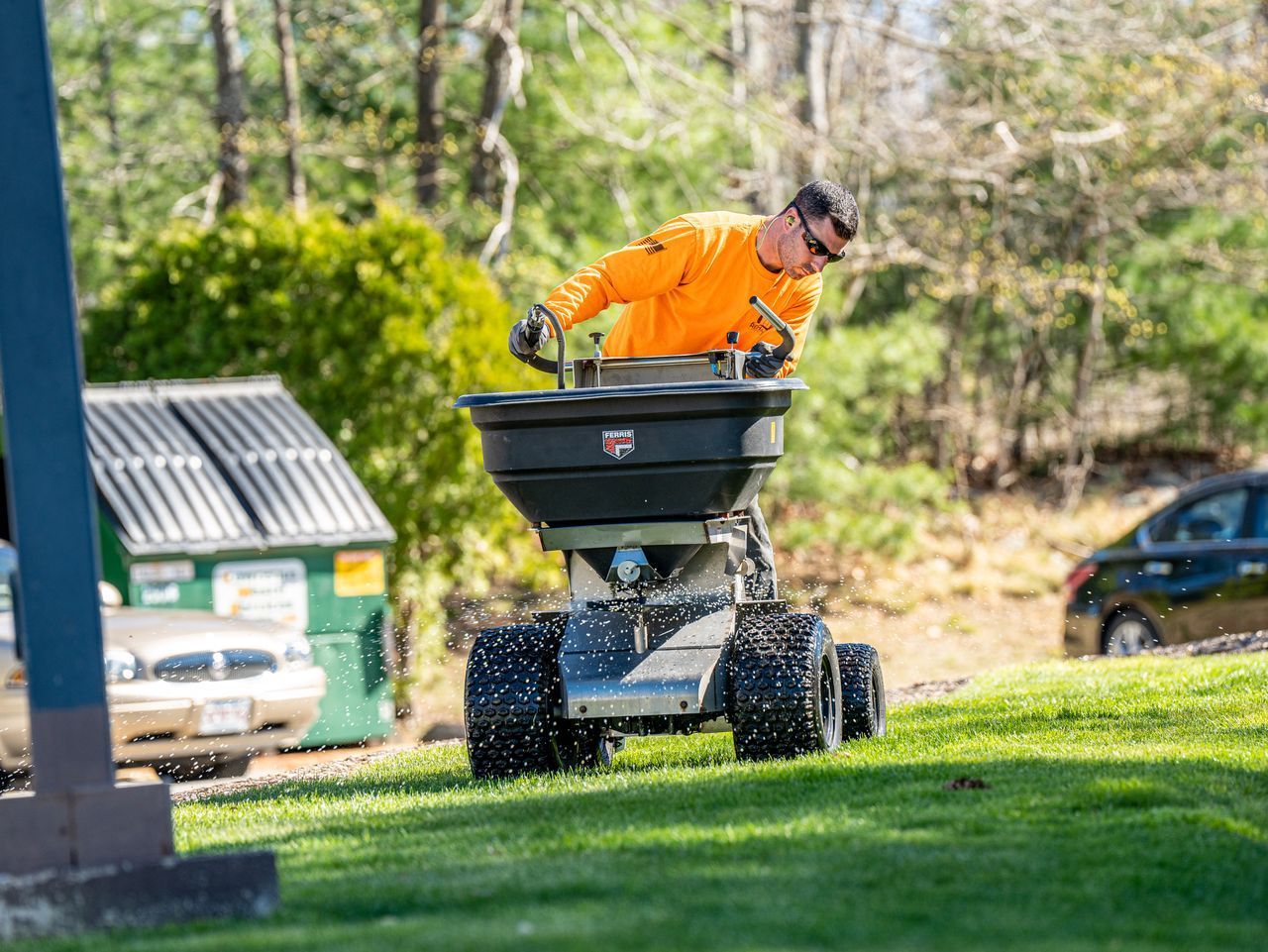 Person in orange work jacket spreading material from a black spreader on a grassy lawn.