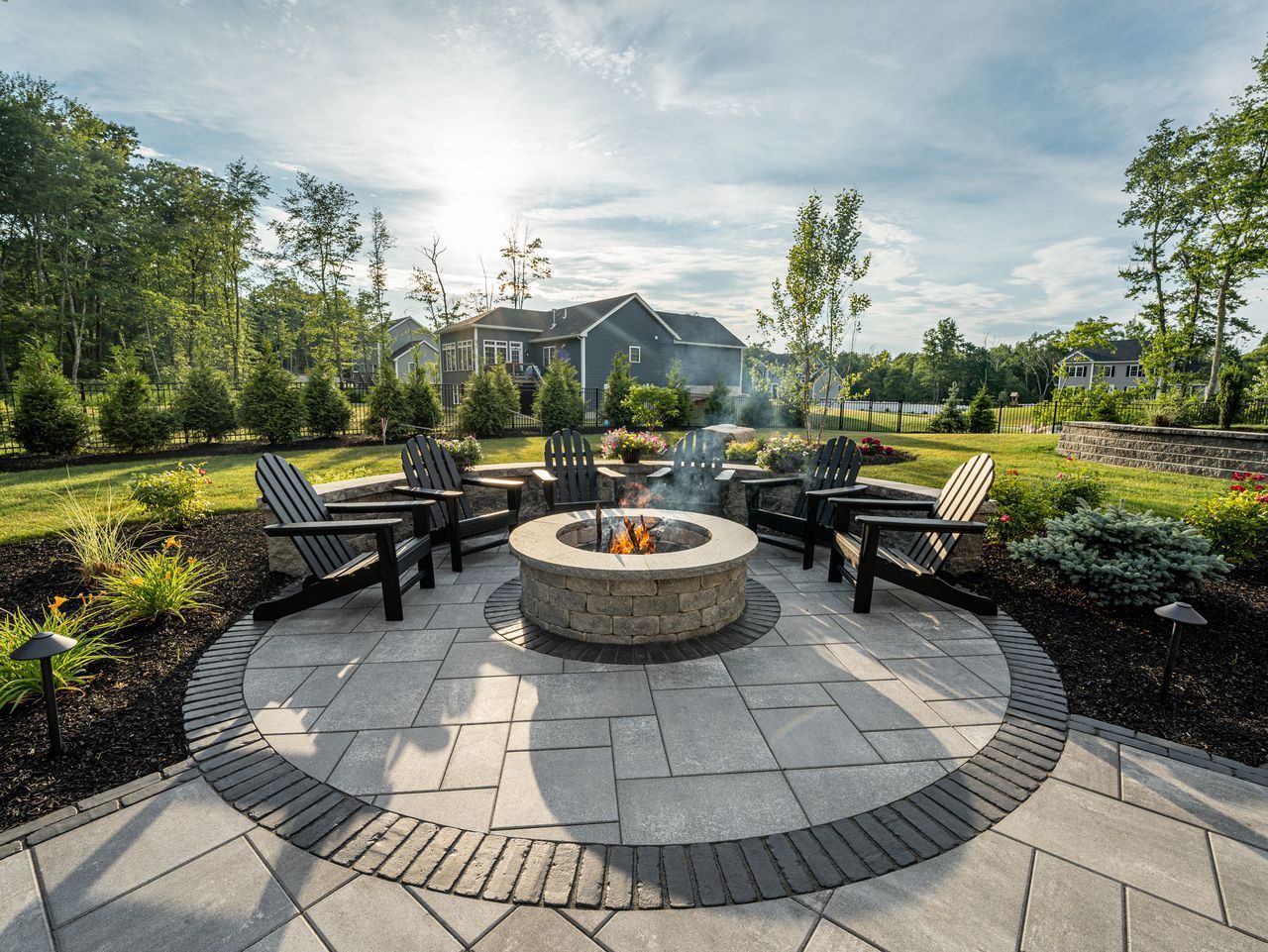 Outdoor fire pit seating area with chairs, stone pavers, and landscaped garden under a sunny sky