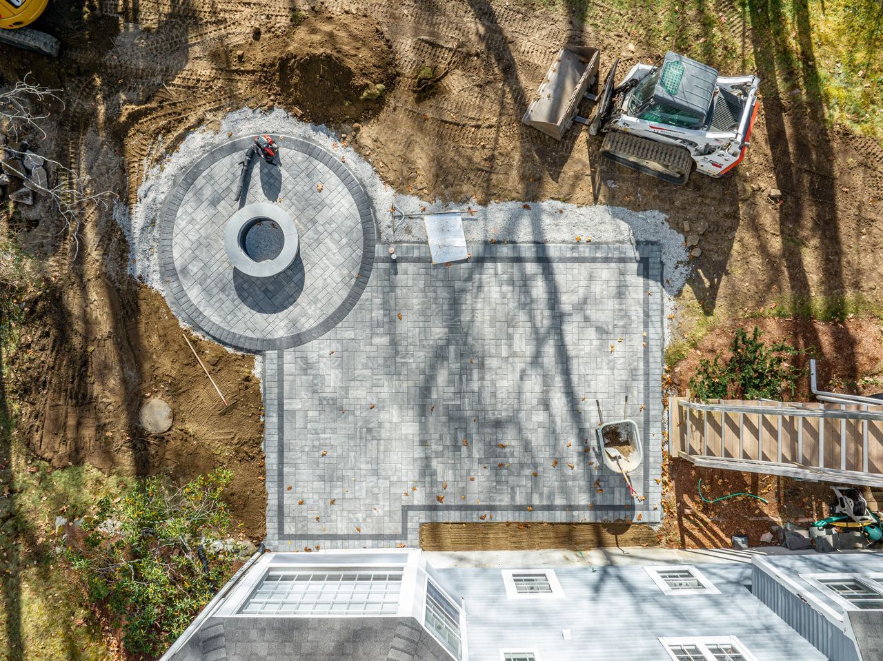 Aerial view of a backyard patio with a circular fire pit and nearby ladder, cast with tree shadows.