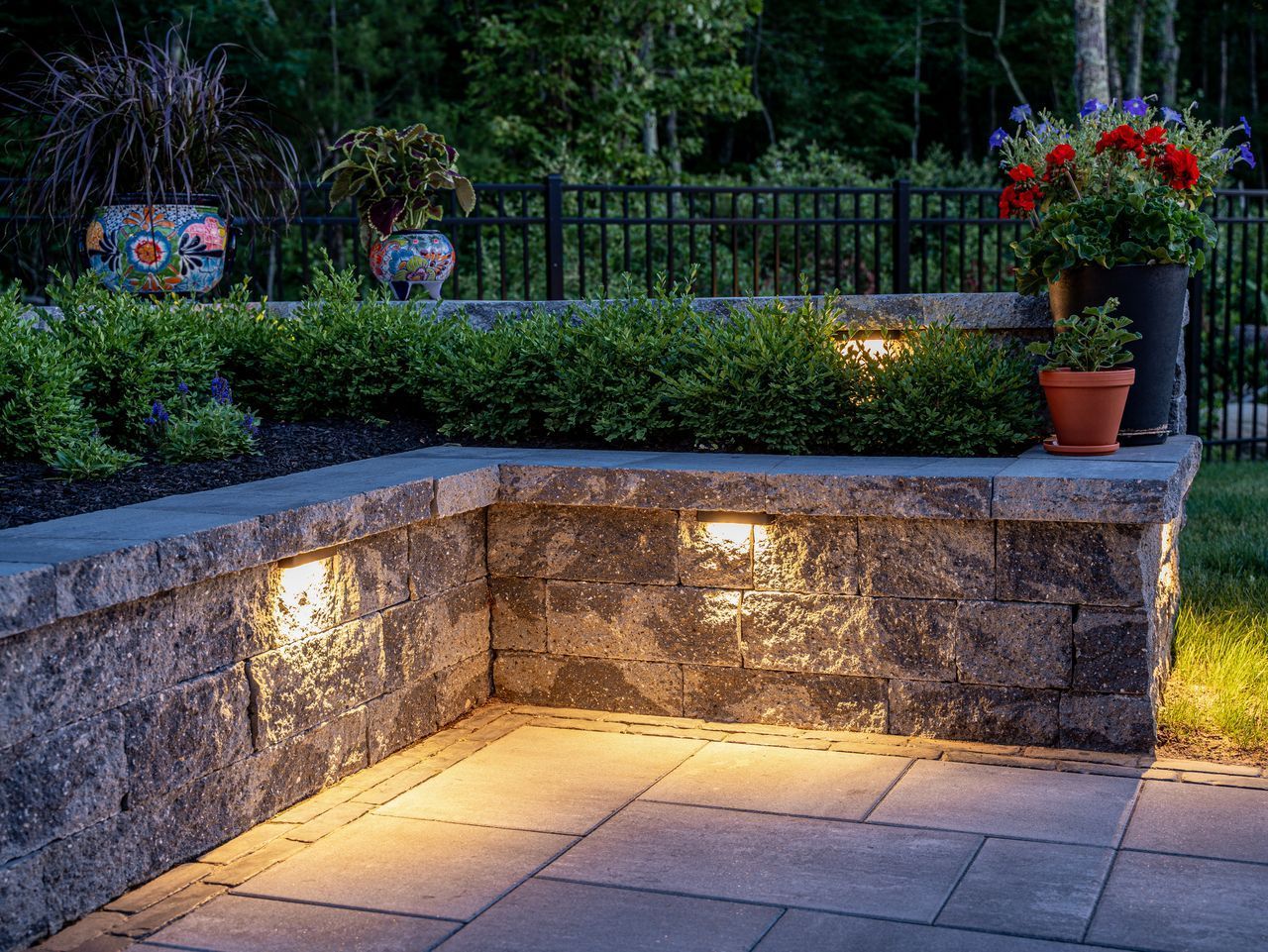 Stone patio corner with warm ground lights, potted red flowers, and a garden wall at dusk
