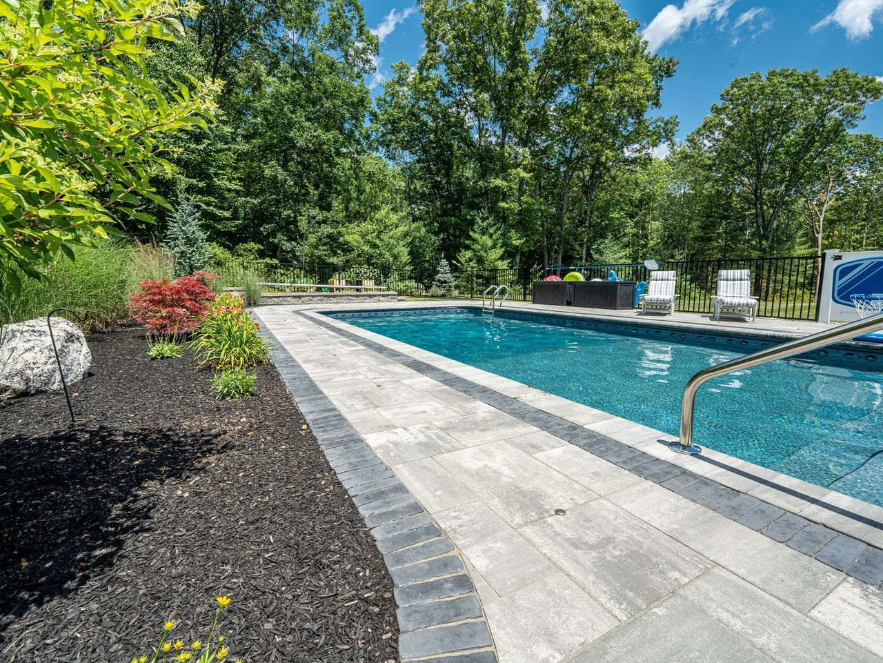 Backyard swimming pool with blue water, stone deck, and lush green trees under a clear sky