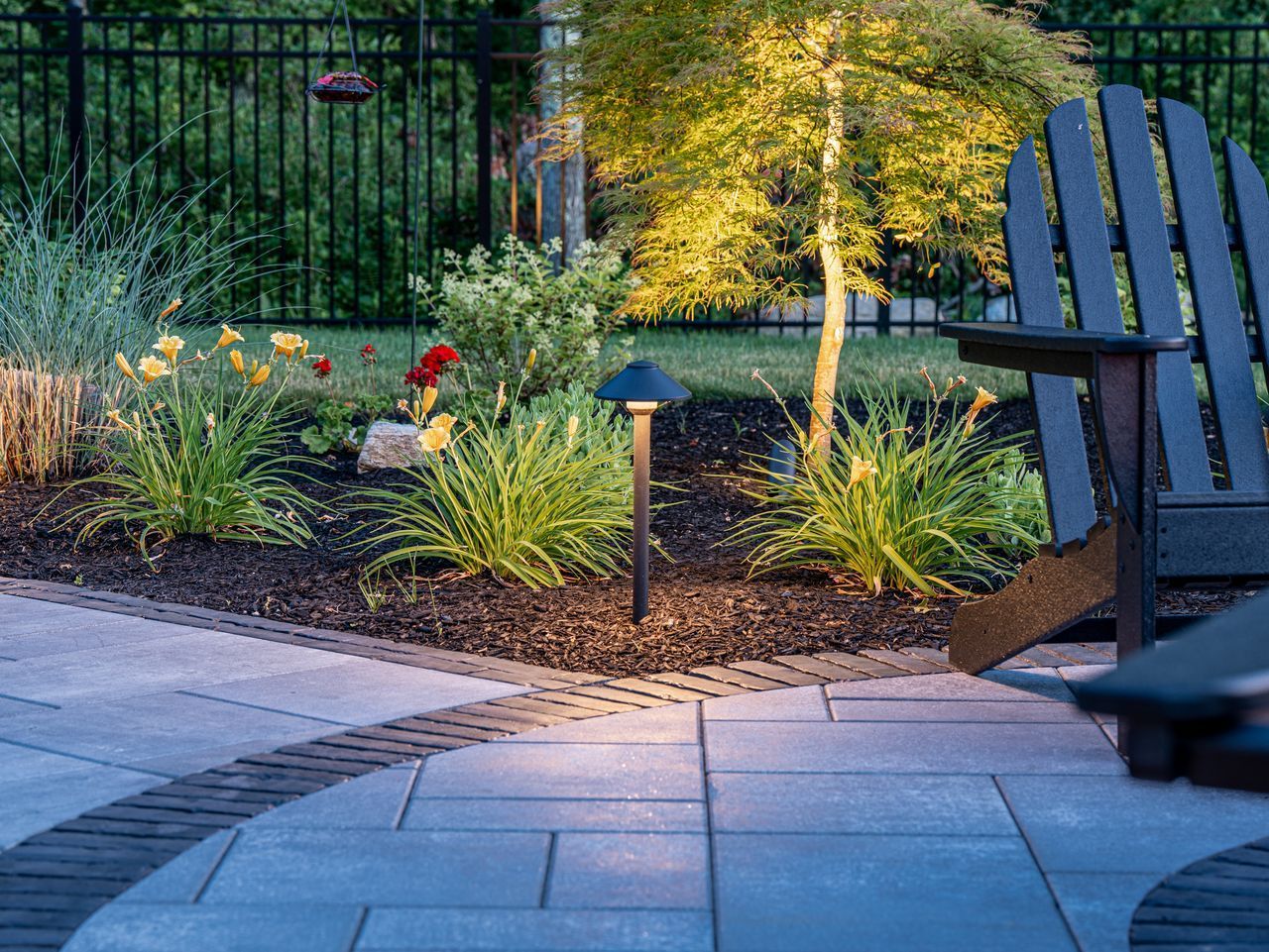 Landscaped backyard patio with stone pavers, lit shrubs, and a dark Adirondack chair by a fence