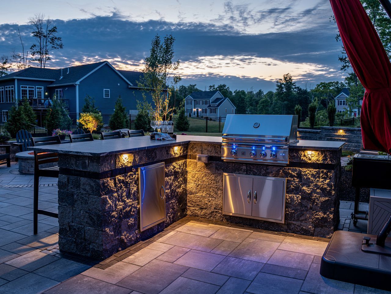 Outdoor patio with lit stone grill and sink at dusk, overlooking a neighborhood and mountains
