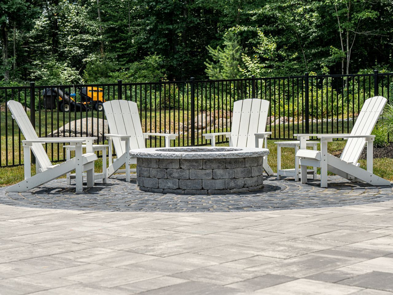 Stone fire pit surrounded by white Adirondack chairs on a patio, with a black fence and trees behind.