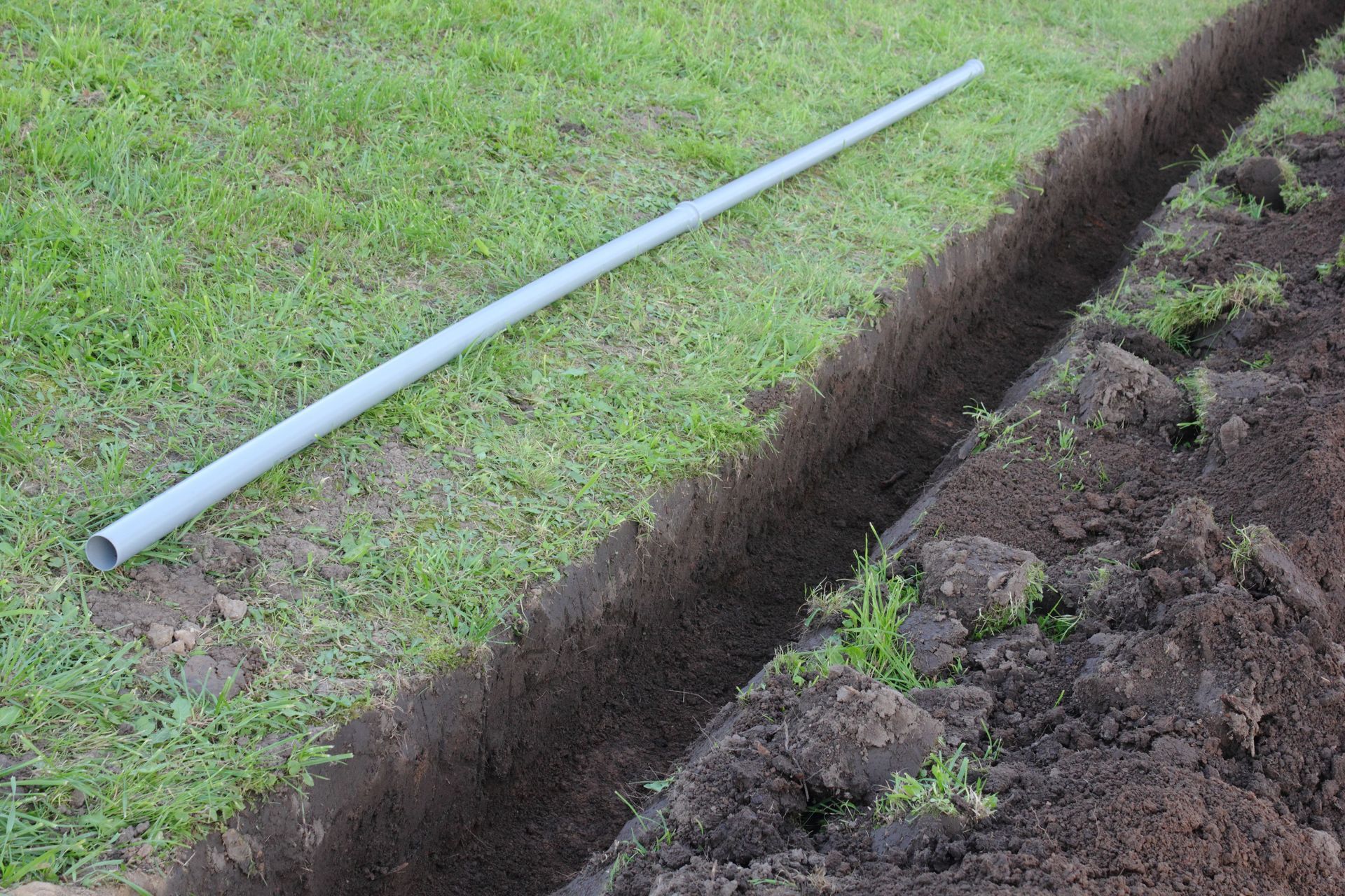 White PVC pipe lying on grass beside a freshly dug trench along a dirt edge.