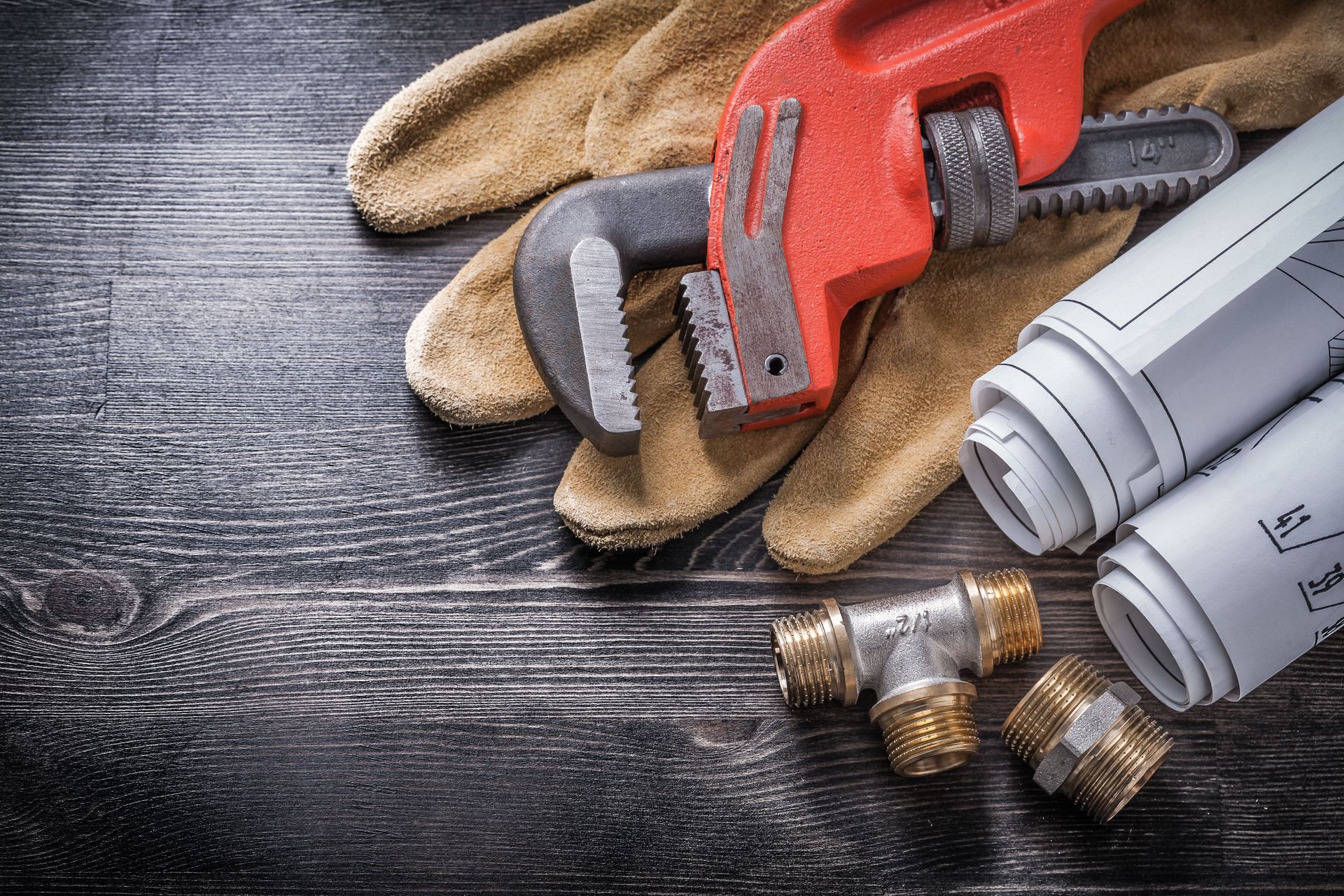 Plumbing tools: wrench, gloves, blueprints, fittings, on a dark wooden surface.