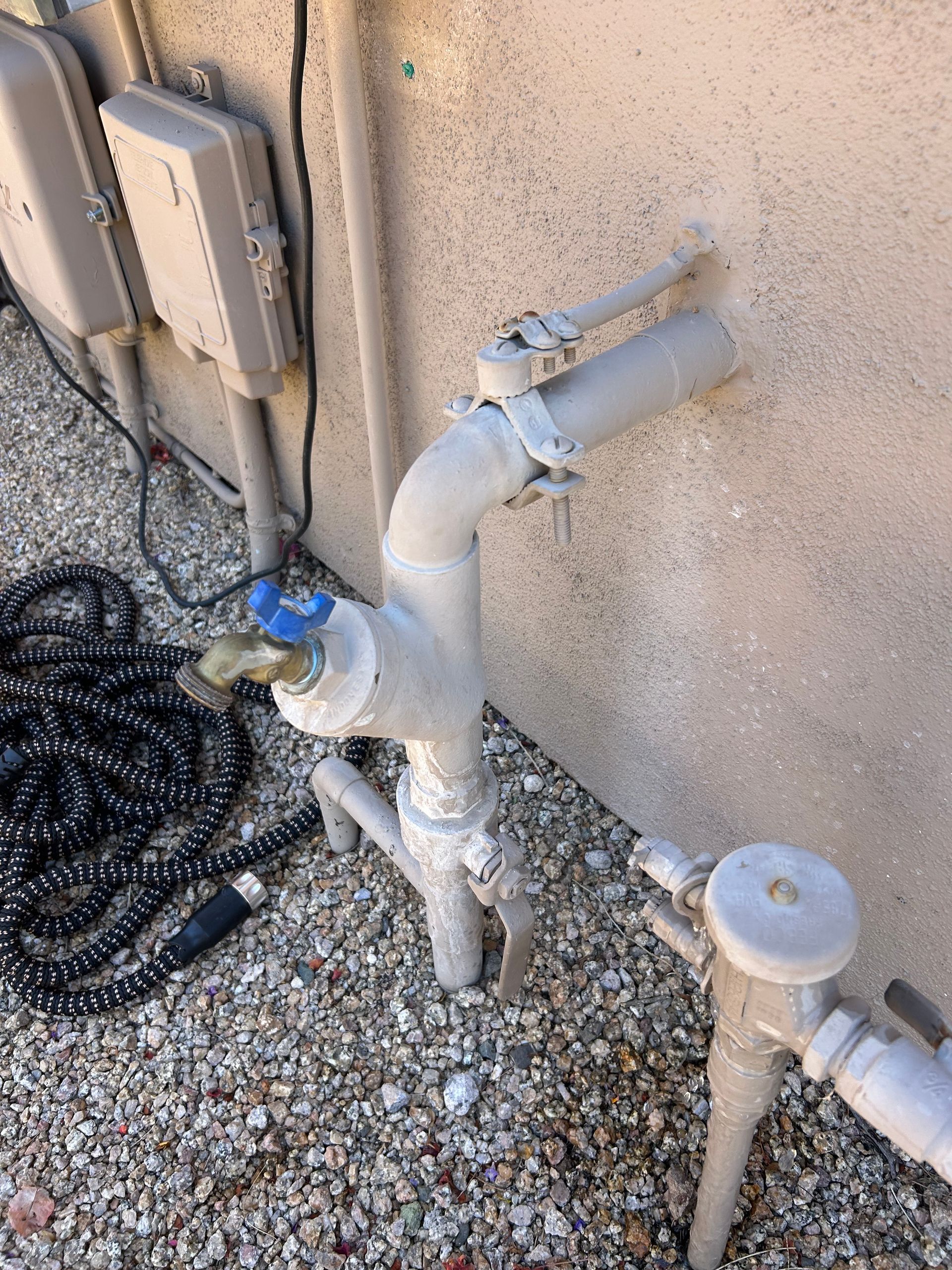 Outdoor water faucet with pipes against a beige wall, next to a coiled black hose.