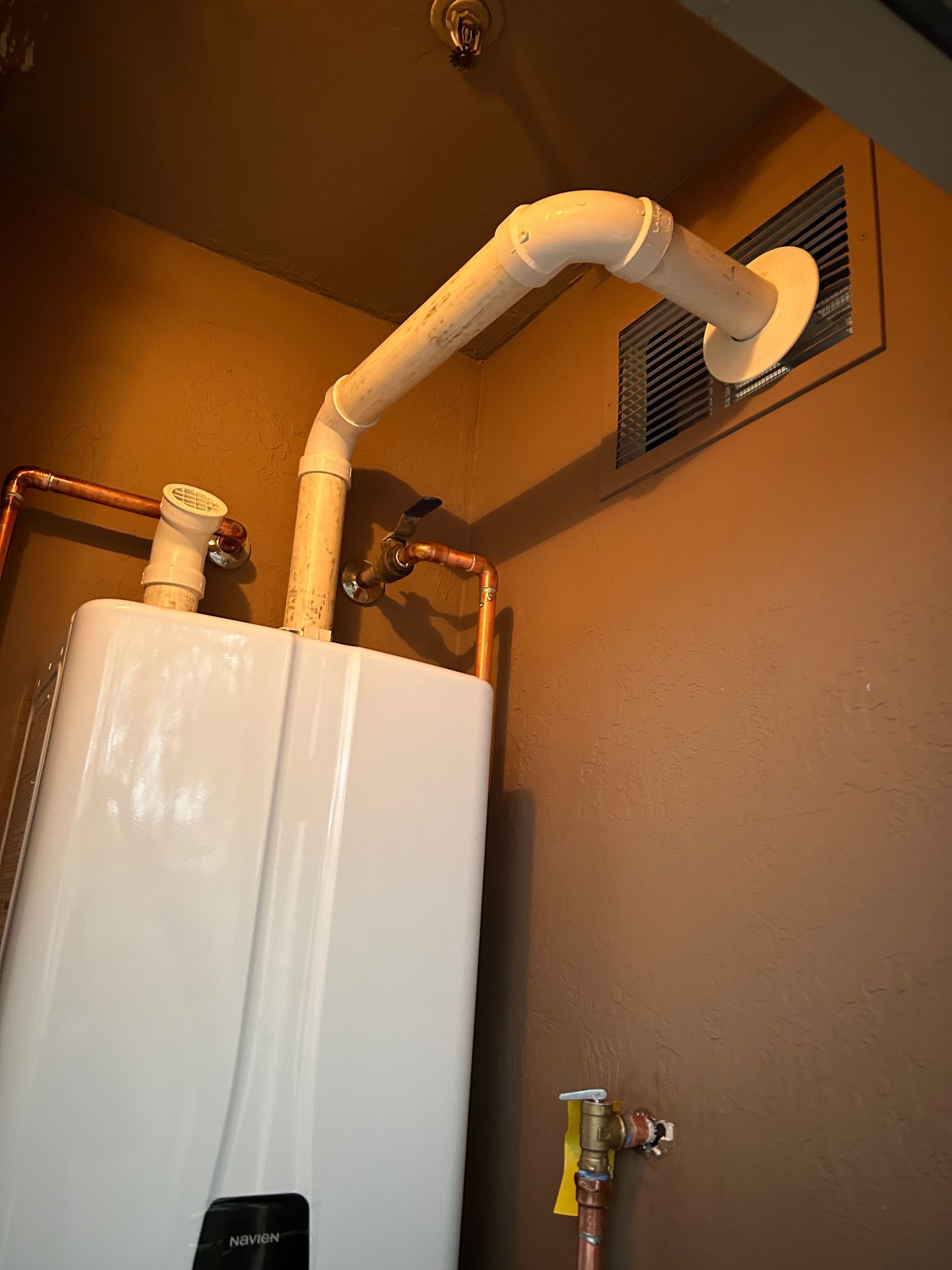 White water heater with white exhaust pipe venting to a wall vent. Brown walls.