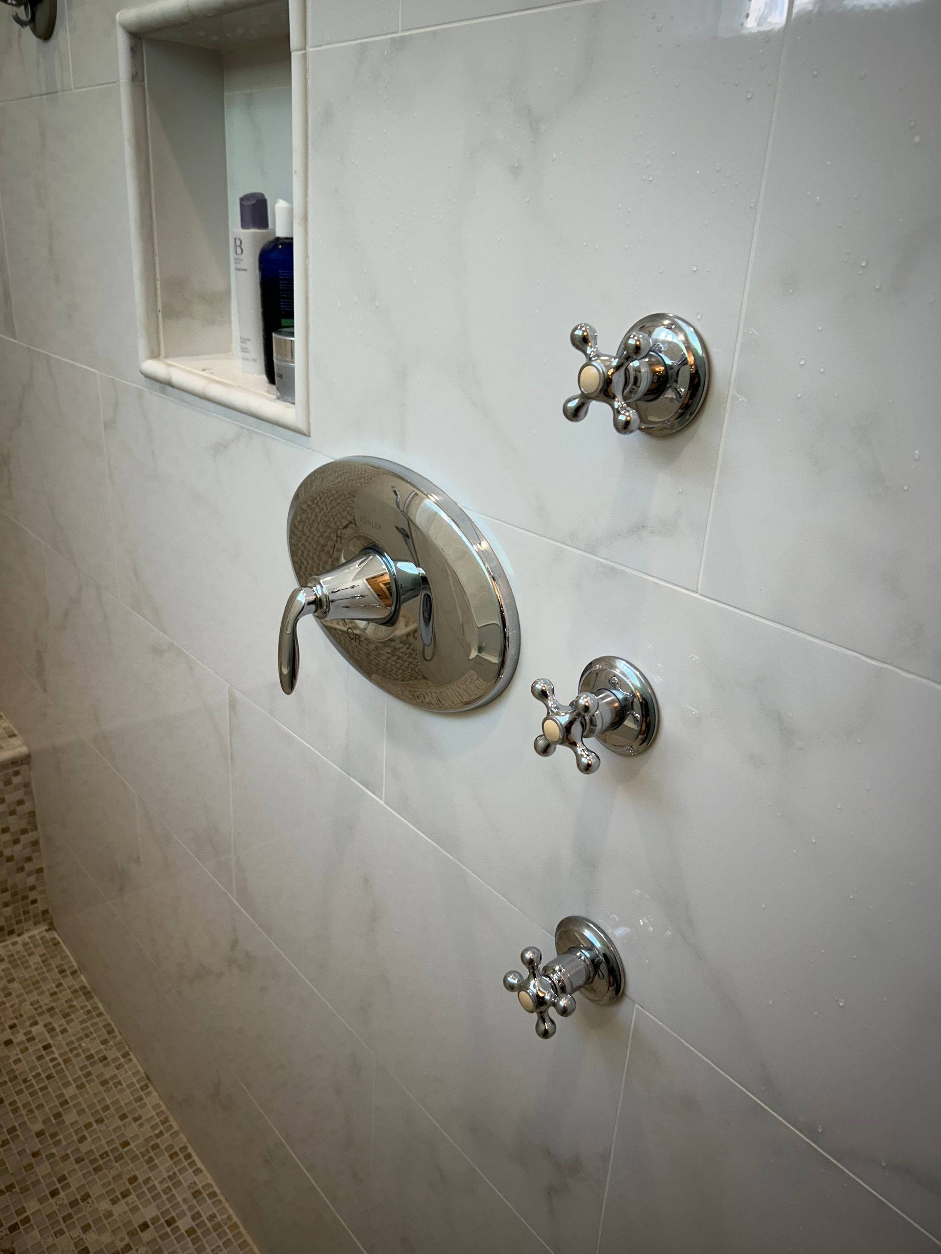 Chrome shower fixtures on a white marble tiled wall with a recessed shelf.