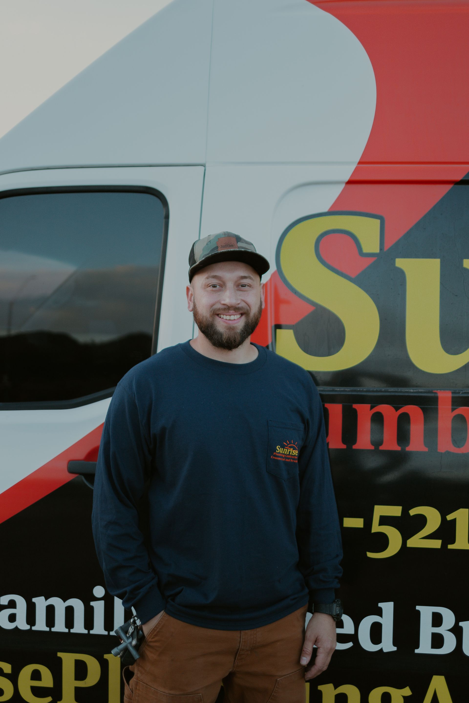 Man in blue shirt stands next to a plumbing van; he wears a hat and smiles.