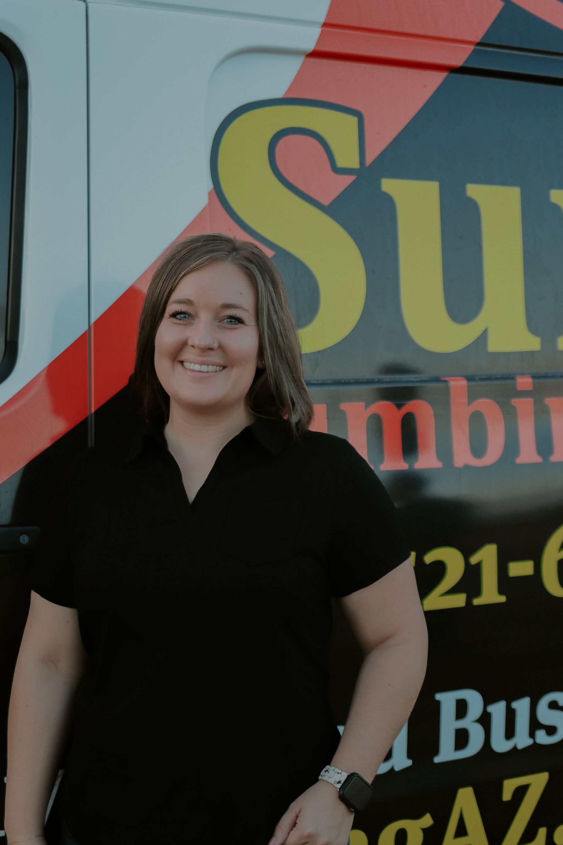 Woman in black shirt smiles, posing by a Sun Plumbing van.