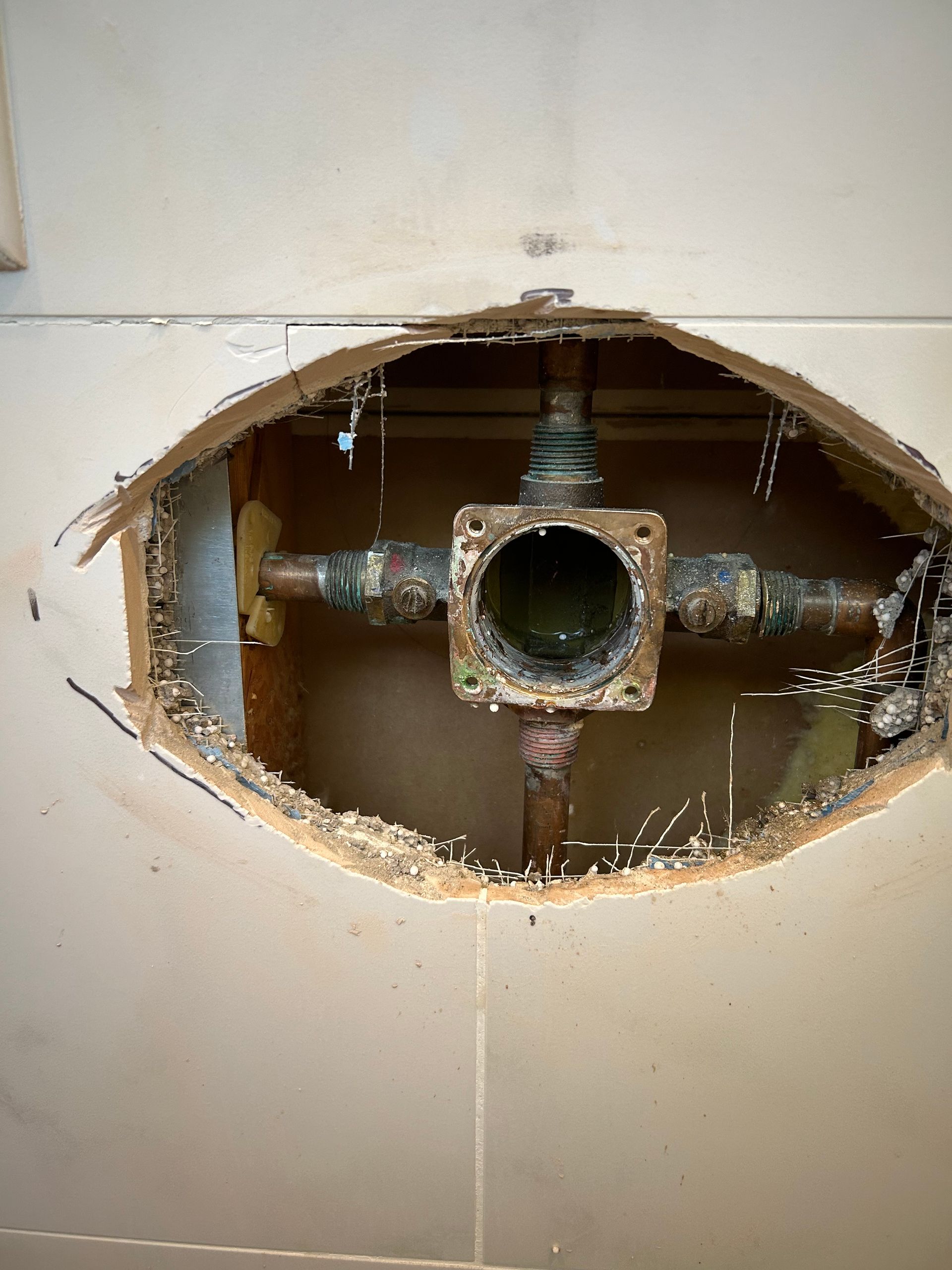 Hole in a wall revealing shower valve and copper pipes, surrounded by damaged tile.