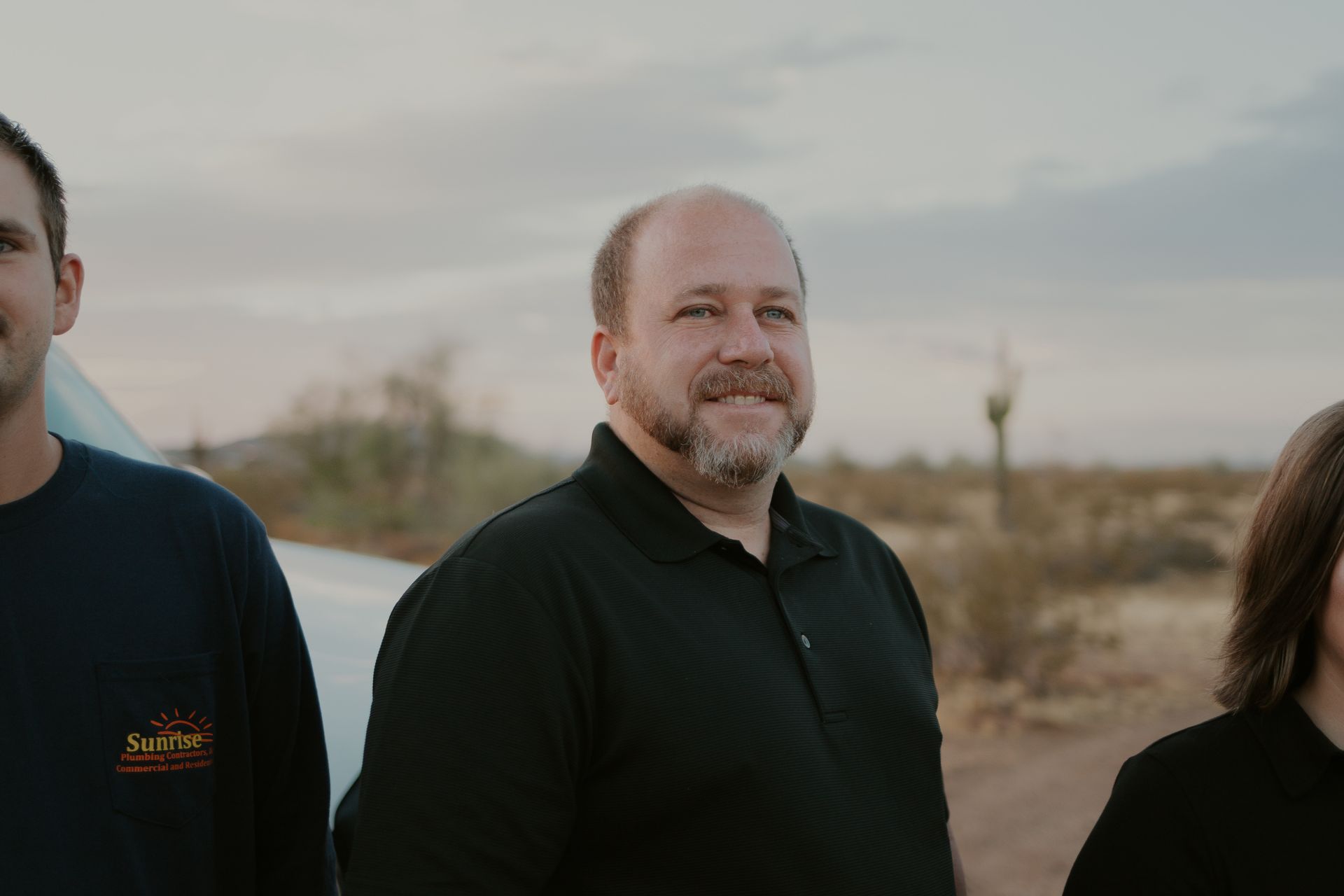 Three people standing in an arid landscape; middle man with beard smiles at camera.